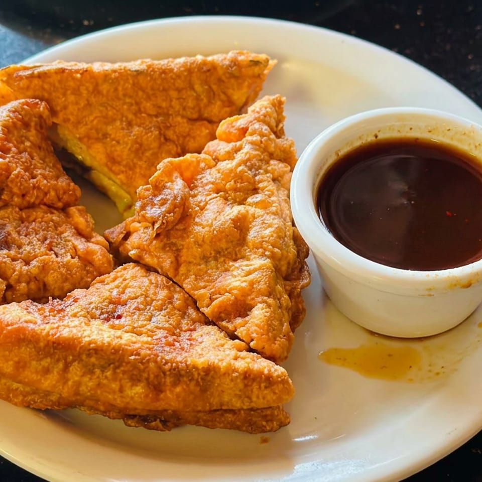 Plate of golden fried sandwiches with a small cup of dark dipping sauce on a dark countertop.
