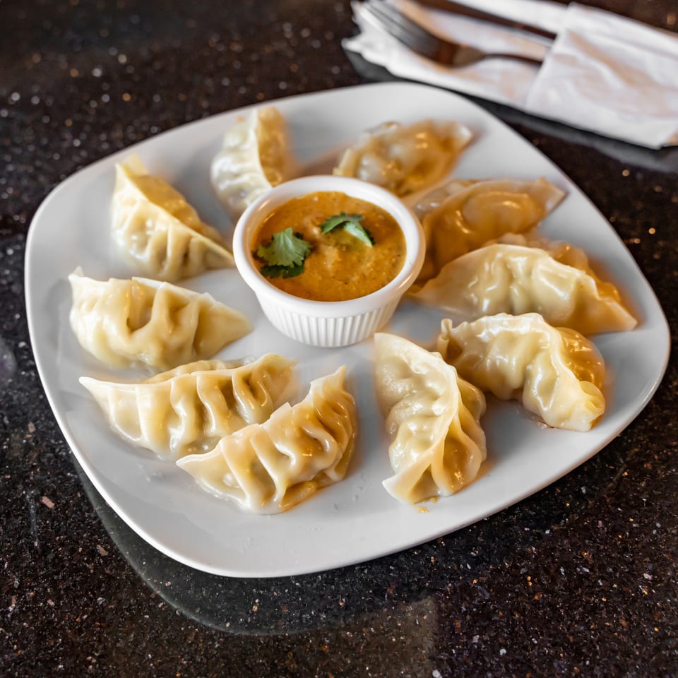 Steamed dumplings arranged around a bowl of cilantro-topped orange dipping sauce on a white plate.
