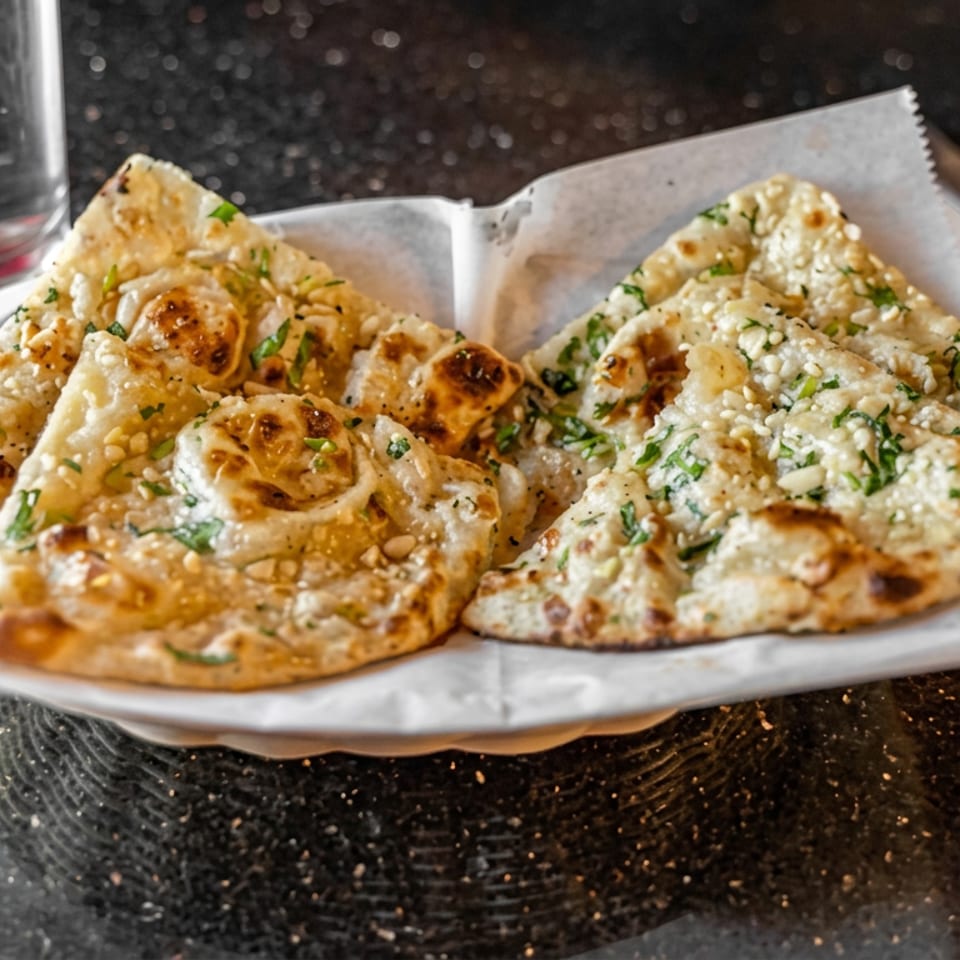 Sliced, herb-topped flatbread with toasted cheese and pine nuts served on parchment over a dark restaurant table.