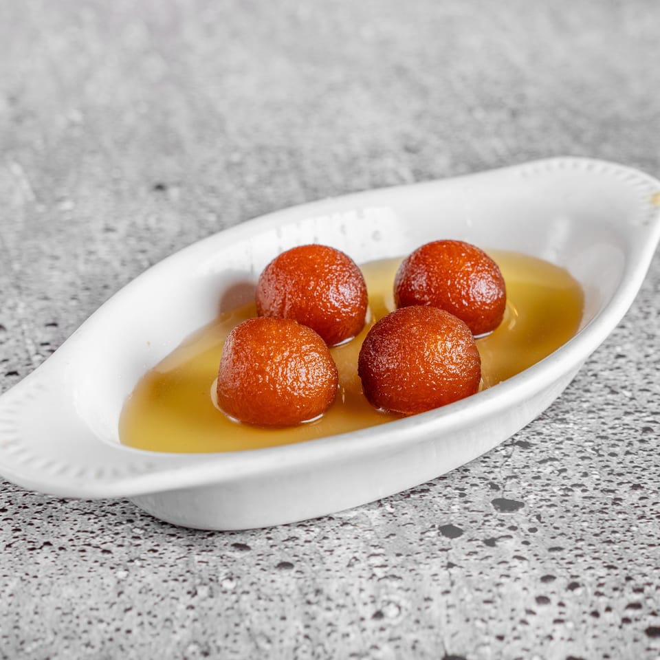 Four golden-brown syrup-soaked dessert balls served in a white oval dish on a speckled countertop.