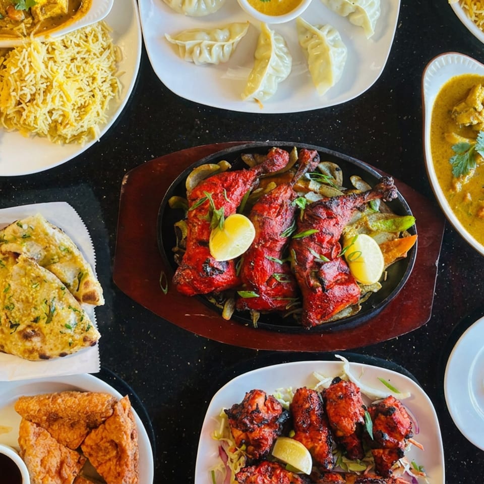 Overhead spread of Indian dishes including biryani, curries, dumplings, naan, samosas, tandoori meats, and orange juice on a table.