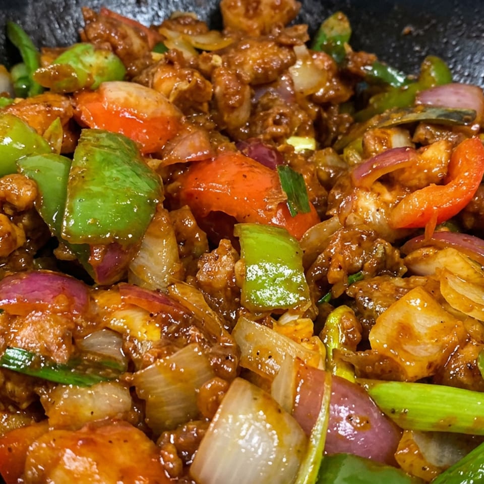 Glossy stir-fried chicken with green and red peppers, onions, tomatoes, and scallions in a black wok.