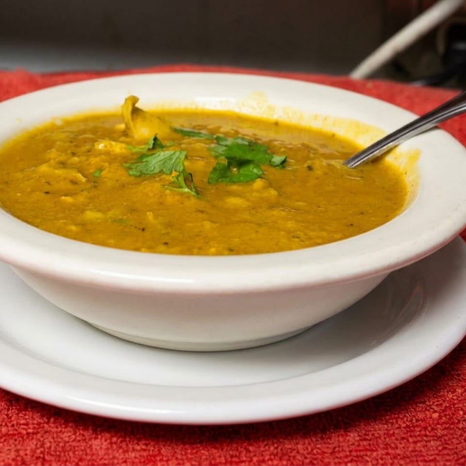 Bowl of golden lentil soup garnished with fresh cilantro, served with a spoon on a white plate atop a red tablecloth.